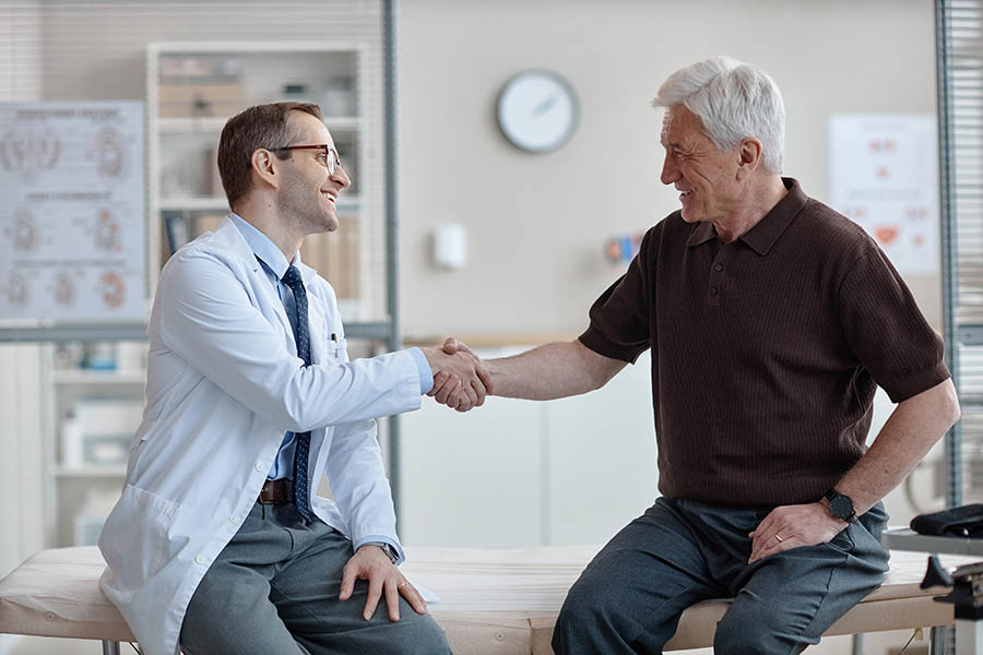 Middle aged Caucasian male doctor wearing glasses shaking hands with senior Caucasian man sitting on examination table in medical office during urology consultation
